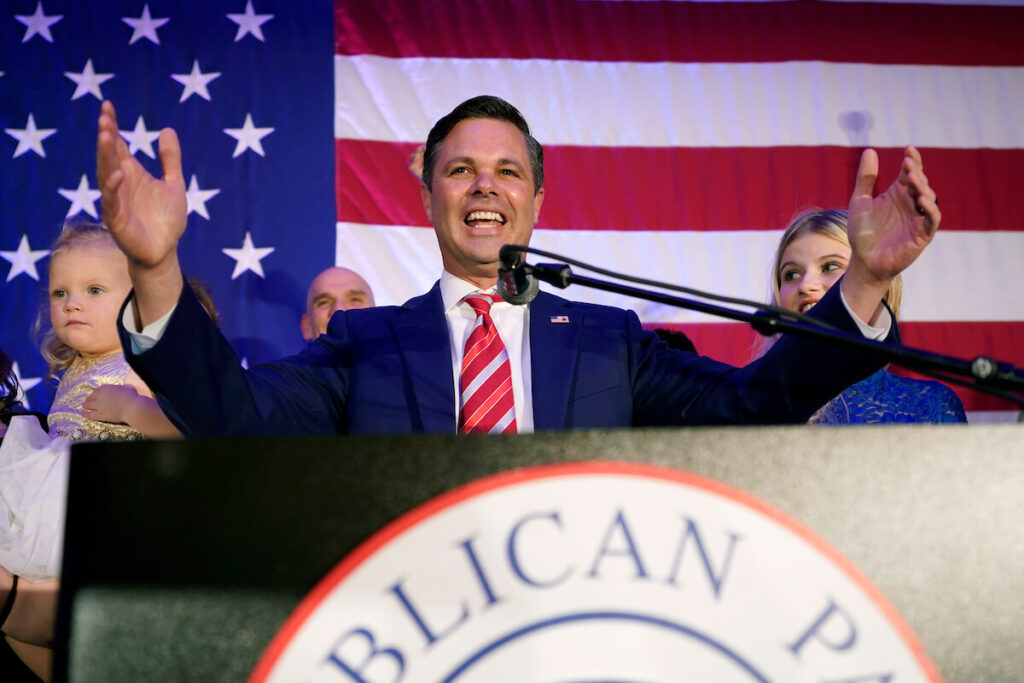 Rep. Zach Nunn stands behind a podium, in front of an American flag during a watch party for the 2022 election. He's smiling and his arms are raised as he gives his victory speech.