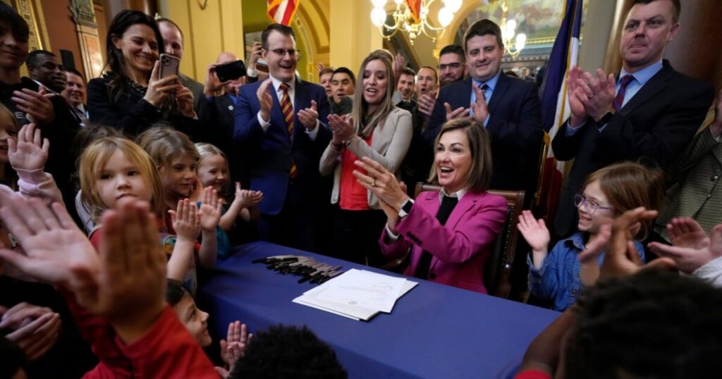 Gov. Kim Reynolds signs the private school voucher bill in front of a crowd of children and lawmakers.