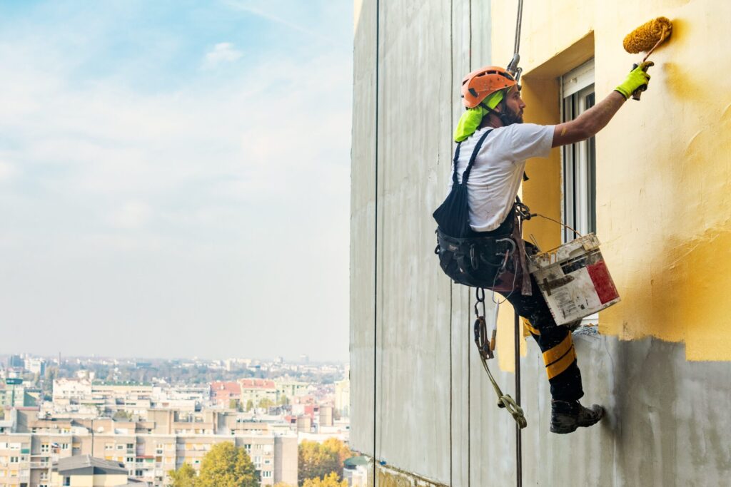 Industrial rope access worker hanging from the building while painting the exterior facade wall. Industrial alpinism concept image.