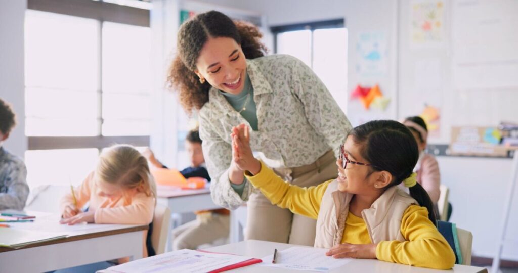 Teacher woman, high five girl and classroom with achievement, success and mentorship for learning. Education, development and students with goals, knowledge and books with celebration at school desk