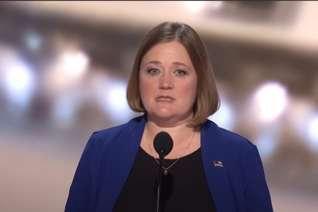 Brenna Bird standing in front of a microphone at the RNC. She has a black shirt with a blue jacket over it.