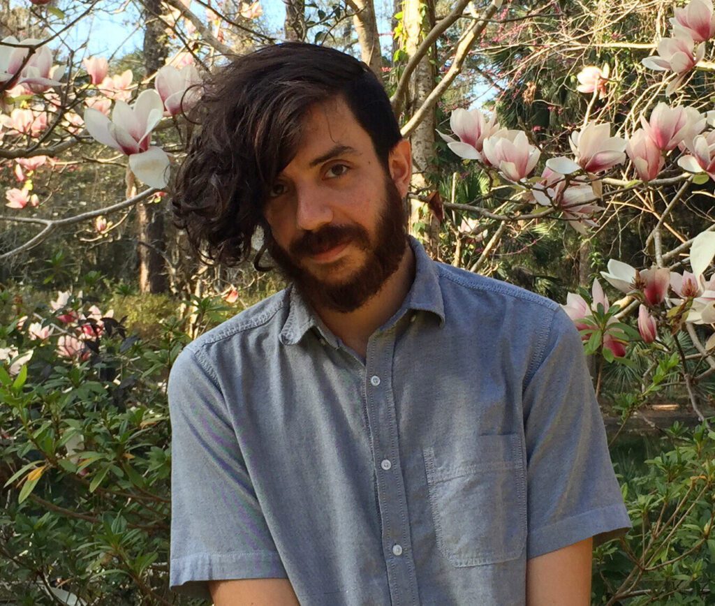 Photo of Kaveh Akbar pictured in a blue shirt outside against a background of trees and flowers.