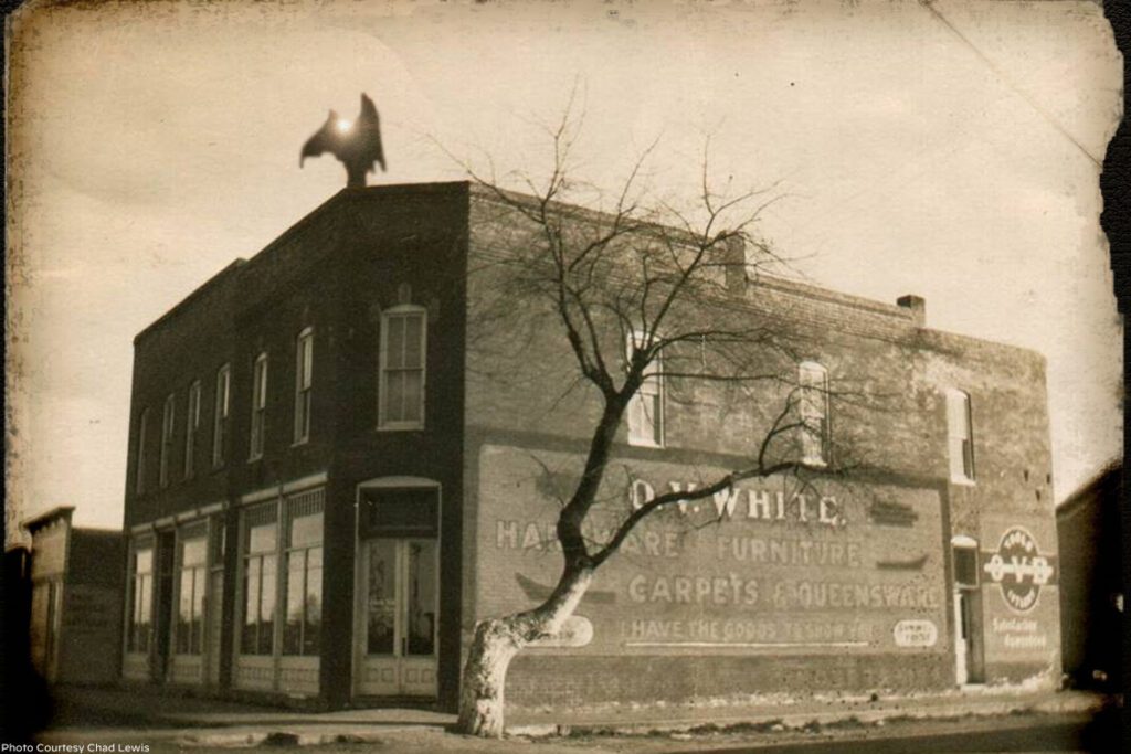 An old sepia-colored photo of an old building with the Van Meter Visitor percehd on top. The Visitor is a large, pointy shape with wings and a bright light shines from its forehead.