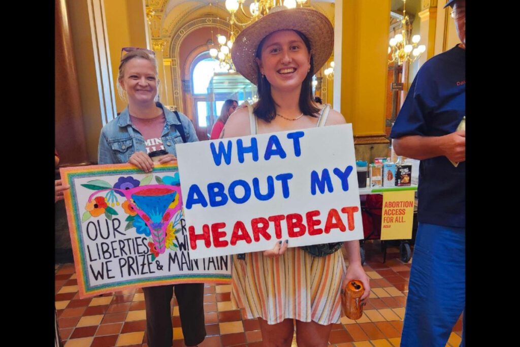 Photo is of signs held by two women at the protest of the 2023 near-total abortion ban. Both are in the Capitol Rotunda. The woman in the center is wearing a hat and smiling over her sign that reads "what about my heartbeat." On the left, just behind her, is a woman in a jean jacket who is also smiling and holding a sign that reads "Our liberties we prize and our rights we will maintain." The words are around an illustration of a uterus that's surrounded by flowers.